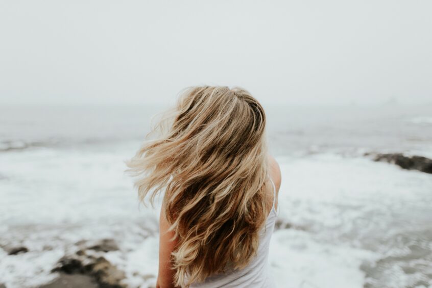 A girl with long blonde hair standing on an overcast beach. Her back is to the camera.