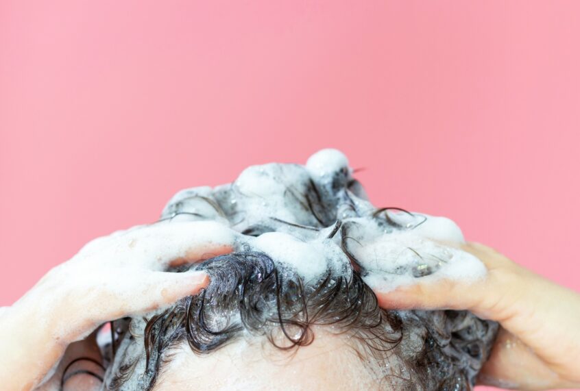 A close-up of a person shampooing their hair. The background is pink.